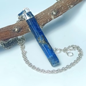 May include: A blue kyanite crystal pendant necklace with a silver chain. The kyanite is a deep blue colour with natural variations. The necklace is displayed on a brown branch against a light blue background. The chain is a delicate silver link.