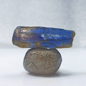 Pu&ograve; includere: Una pietra preziosa blu e dorata con una texture ruvida e naturale. La pietra preziosa &egrave; in equilibrio su un piccolo sasso rotondo grigio.