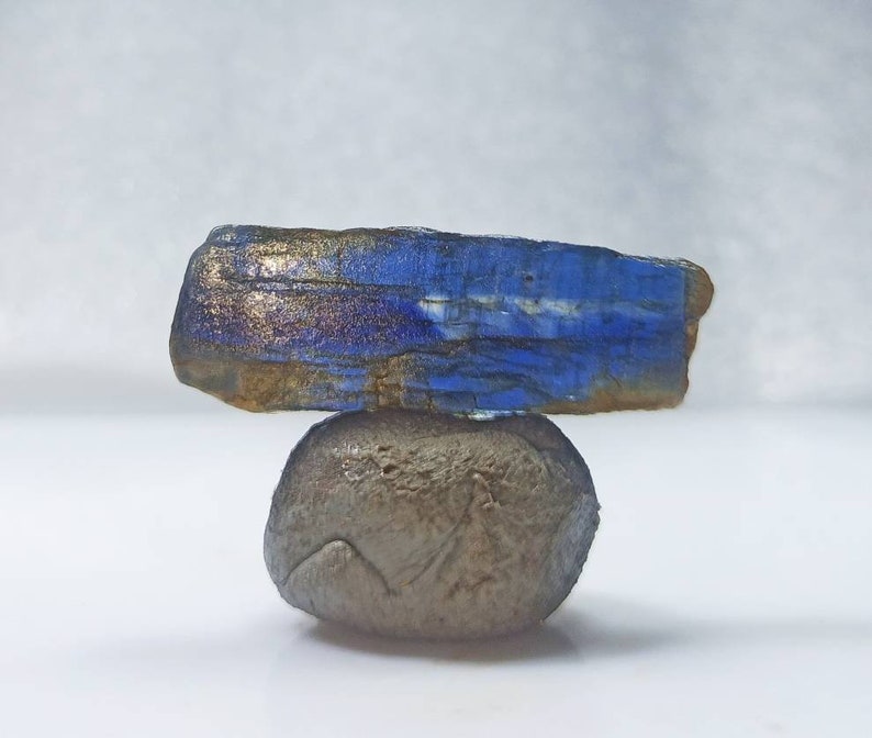 Pu&ograve; includere: Una pietra preziosa blu e dorata con una texture ruvida e naturale. La pietra preziosa &egrave; in equilibrio su un piccolo sasso rotondo grigio.