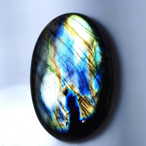 Puede incluir: Una piedra preciosa de labradorita de forma ovalada con una superficie pulida. La piedra muestra un vibrante juego de colores, con destellos iridiscentes de azul, verde y amarillo. La piedra está sobre un fondo blanco.