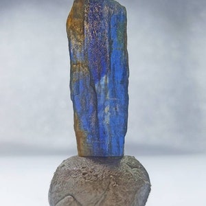Pu&ograve; includere: Un cristallo blu e oro su una roccia grigia. Il cristallo &egrave; alto e sottile, con una texture ruvida. La roccia &egrave; rotonda e liscia.