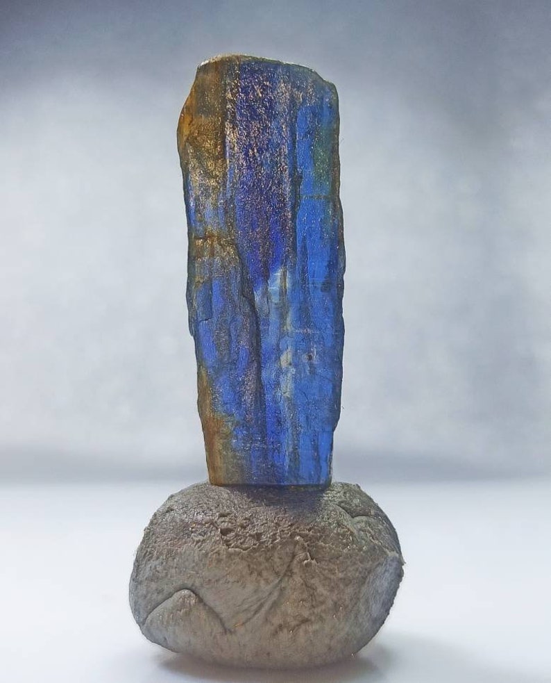 Pu&ograve; includere: Un cristallo blu e oro su una roccia grigia. Il cristallo &egrave; alto e sottile, con una texture ruvida. La roccia &egrave; rotonda e liscia.
