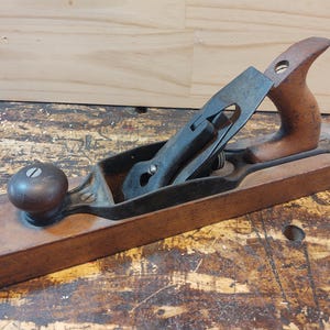 May include: A vintage wooden hand plane with a black metal blade and a wooden handle. The plane is sitting on a wooden surface.