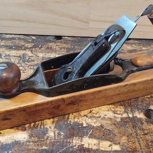May include: A wooden hand plane with a black metal blade and a wooden handle. The plane is in good condition and has a natural wood finish.