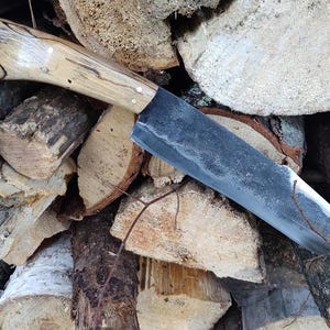 May include: A handcrafted knife with a light-colored wooden handle and a dark, textured blade. The handle is secured with small metal rivets. The knife is displayed against a backdrop of stacked firewood, showcasing its rustic aesthetic.