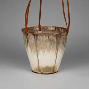 May include: A hanging ceramic planter with a cream and brown glaze. The planter has a flared shape and is suspended by brown leather straps. The glaze has a dripped effect, creating a unique texture.