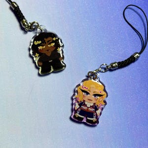May include: Two acrylic charms with cartoon character designs. One charm shows a dark-skinned character in black, the other a blonde character in navy and red. Both have gold accents and are attached to black cords.