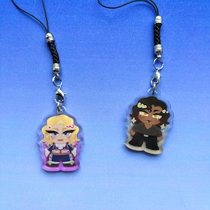 Pode incluir: Dois pingentes de acrílico presos a cordões trançados pretos com fechos de metal prateado. Um pingente apresenta um personagem loiro com roupa azul e vermelha, enquanto o outro retrata um personagem de cabelos escuros com camisa e calça pretas.