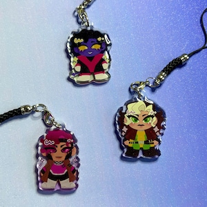 Pode incluir: Três pingentes de acrílico com personagens de desenhos animados. Cada pingente tem um fecho prateado e um cordão preto. Os personagens têm designs únicos com cores vibrantes, incluindo roxo, vermelho, marrom e verde. Os pingentes estão em um fundo azul gradiente.