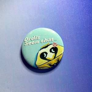 Puede incluir: Un botón redondo azul claro con el texto "Ordis Seen that..." en blanco. El botón presenta un personaje de dibujos animados amarillo y negro con una expresión de enfado. El fondo es un degradado de azul y morado.