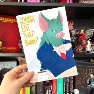 May include: A greeting card featuring a bat-pig hybrid character in a blue suit and red tie, pointing. The card has the text "GONNA EAT THAT TWINK?" in orange. A coffee mug with the text "IT WAS A MISINPUT" is also visible.