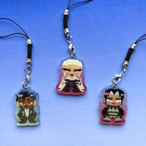 May include: Three acrylic charms featuring stylized cartoon characters, each attached to a black braided cord with a silver metal clasp. The left charm depicts a character with dark hair, the center a blonde character in a white headpiece, and the right a dark-haired character with white markings.