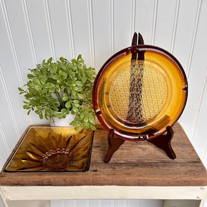 May include: Two amber glass ashtrays with a textured design. The larger ashtray is round and the smaller ashtray is square. Both ashtrays are on a wooden table with a white painted base.