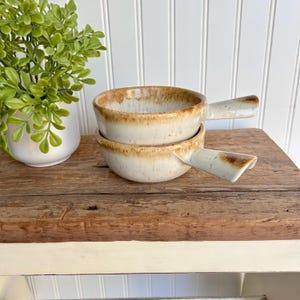 May include: Two white ceramic soup bowls with brown speckled glaze and a handle. The bowls are stacked on top of each other.