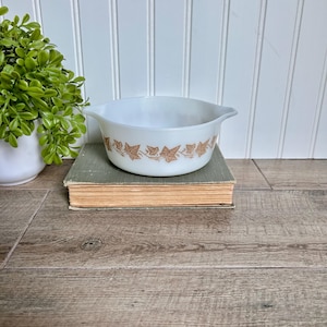 May include: A white Pyrex bowl with a brown ivy leaf pattern around the rim. The bowl is on a vintage book with a green cover. A small green plant in a white pot is in the background.