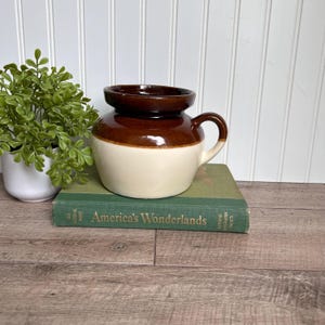 Könnte beinhalten: Ein brauner und weißer Keramiktopf mit einem breiten Rand und einem einzelnen Henkel. Der Topf steht auf einem grünen Buch mit dem Titel "America's Wonderlands".