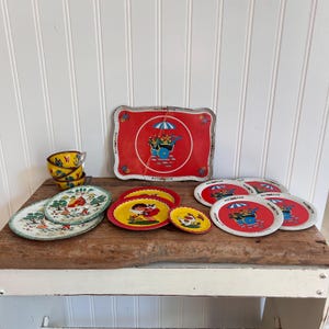 Puede incluir: Una colección de artículos de hojalata vintage con una bandeja roja con un diseño de carro floral, junto con platos y tazas a juego. Los platos y tazas tienen varias ilustraciones, incluyendo una niña, un pollo y una casa. Los artículos están dispuestos sobre una superficie de madera rústica.