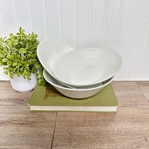 May include: Three white ceramic bowls with a fluted design are stacked on top of each other. The bowls are sitting on a green book.