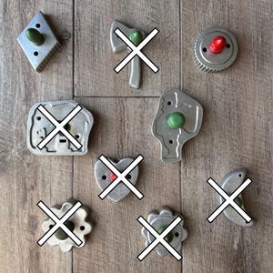 Puede incluir: Una colección de cortadores de galletas de metal vintage en varias formas, incluyendo un diamante, un engranaje y un perfil. Cada cortador tiene un pequeño botón de color. Los cortadores están dispuestos sobre una superficie de madera.