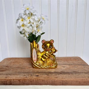 以下が含まれることがあります： クマが小さなクマの子供を抱いている形の金色のセラミックの花瓶。花瓶には側面に温度計があり、白い花が花瓶に入っています。