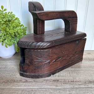 Puede incluir: Una caja de madera vintage con forma de plancha. La caja es de color marrón oscuro con un diseño tallado en la parte delantera. La caja tiene una tapa con bisagras y un asa en la parte superior.