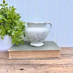 May include: A white ceramic pitcher with a blue floral design. The pitcher has a handle and a rounded base. It is sitting on a book.