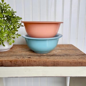 May include: Two vintage mixing bowls, one peach and one light blue, stacked on a wooden surface.