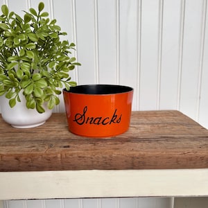 Puede incluir: Tazón de aperitivos de cerámica naranja con borde negro y la palabra "Snacks" escrita en letras cursivas negras.