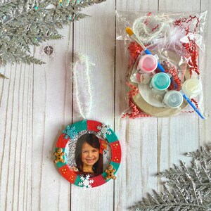 May include: A red and green Christmas ornament with a photo frame, gingerbread men, and snowflakes. The ornament is decorated with glitter and comes with a white string for hanging. The ornament is packaged with paint, a paintbrush, and a small bag of glitter.