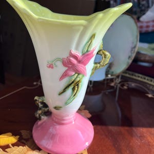May include: A vintage ceramic vase with a flared top and a pink base. The vase features a white body with a pink flower and green leaves. The top is a pale yellow color. A gold handle is on the side.