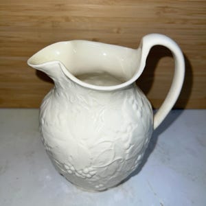 May include: Off-white ceramic pitcher with a curved handle and a spout. The pitcher features a raised relief pattern of leaves and grapes, adding a decorative touch. The pitcher is likely used for serving beverages.