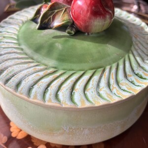 May include: A ceramic serving dish with a lid, featuring a green base and lid. The lid is adorned with a sculpted border and a decorative apple and leaf motif. The dish has a diameter of approximately 8 inches.