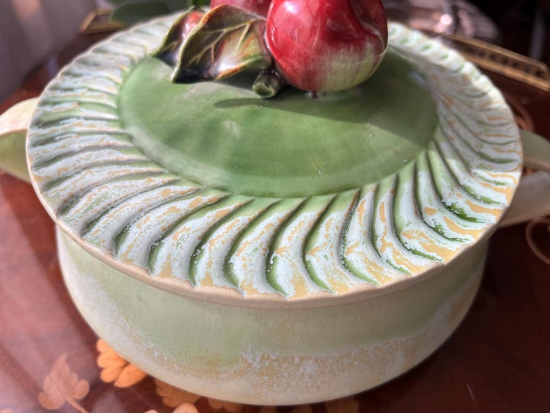 May include: A ceramic serving dish with a lid, featuring a green base and lid. The lid is adorned with a sculpted border and a decorative apple and leaf motif. The dish has a diameter of approximately 8 inches.