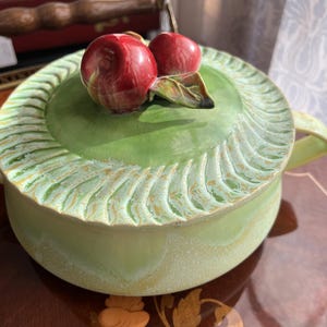 May include: A ceramic container with a lid, featuring a light green base and lid with a textured, ridged design. The lid is topped with two red, glossy cherry sculptures and green leaves. The container has two curved handles.