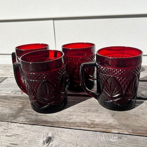 May include: A set of four ruby red glass mugs featuring a pressed diamond pattern on the upper section and a leaf-like motif on the lower body. Each mug has a sturdy handle and a smooth rim, showcasing a deep, translucent crimson color.