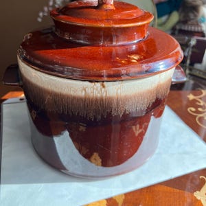 May include: A vintage, two-toned brown ceramic casserole dish with a lid. The dish has a round shape with a handle on the lid and two side handles. The color transitions from a light beige to a dark brown.