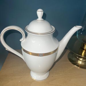 May include: White porcelain teapot with a gold band and accents. The teapot has a curved handle and spout, and a lid with a small knob. The design is simple and elegant, suitable for serving tea. The teapot is on a wooden surface.