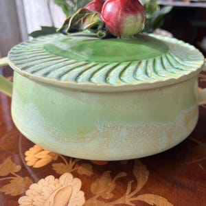 May include: A light green ceramic serving bowl with a matching lid. The lid is decorated with a green leaf design and topped with red apple-shaped ornaments. The bowl has two handles and a textured rim.