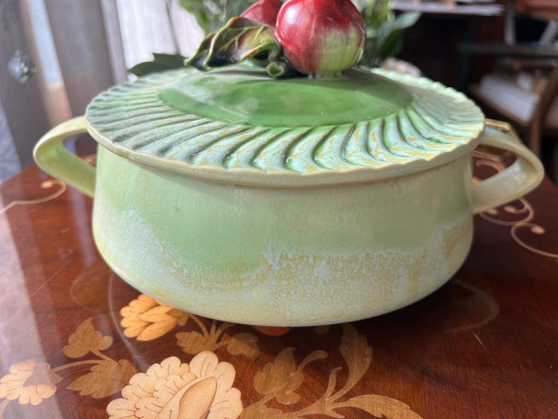 May include: A light green ceramic serving bowl with a matching lid. The lid is decorated with a green leaf design and topped with red apple-shaped ornaments. The bowl has two handles and a textured rim.