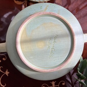 May include: A light green ceramic plate with two handles, viewed from the underside. The plate has a circular design with a raised rim in a lighter shade of green. The text "Made in Calif. U.S.A." is stamped on the plate.