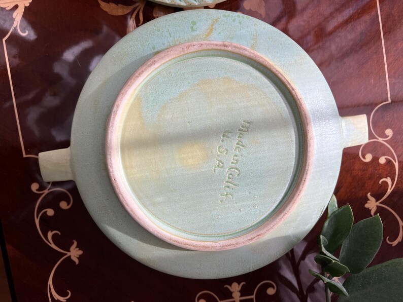 May include: A light green ceramic plate with two handles, viewed from the underside. The plate has a circular design with a raised rim in a lighter shade of green. The text "Made in Calif. U.S.A." is stamped on the plate.