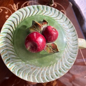 May include: A decorative ceramic plate with a green center and a white and gold patterned rim. Two red apples with green leaves are in the center. The plate has two handles.