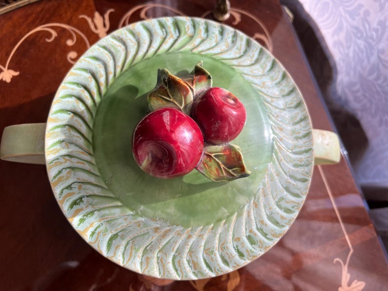 May include: A decorative ceramic plate with a green center and a white and gold patterned rim. Two red apples with green leaves are in the center. The plate has two handles.
