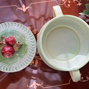 May include: A light green ceramic serving bowl with two handles and a matching plate. The plate features a raised design of two red apples with green and brown leaves. The bowl and plate are on a dark brown surface.
