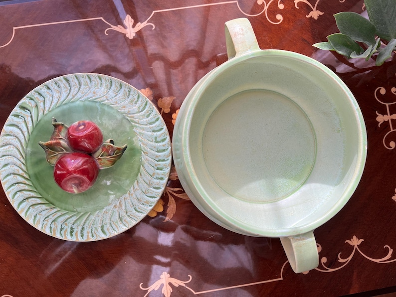May include: A light green ceramic serving bowl with two handles and a matching plate. The plate features a raised design of two red apples with green and brown leaves. The bowl and plate are on a dark brown surface.