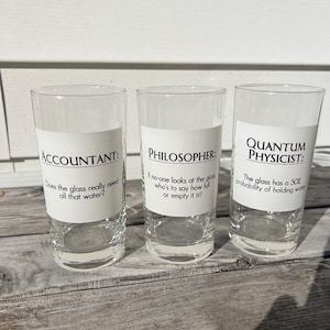 May include: Three clear drinking glasses, each featuring a white label with black text. The first reads: ACCOUNTANT: Does the glass really need all that water? The second: PHILOSOPHER: If no-one looks at the glass who's to say how full or empty it is? The third: QUANTUM PHYSICIST: The glass has a 50% probability of holding water.