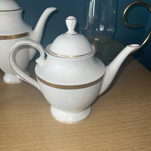May include: White porcelain teapot with a gold band and trim. The teapot has a curved handle and a spout. A matching lid with a small knob sits on top. Another teapot is partially visible in the background. The teapots are on a wooden surface.