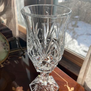 May include: A clear, cut-glass vase with a flared top and a geometric design. The vase sits on a hexagonal base and features intricate patterns throughout. The vase is approximately 8 inches tall.