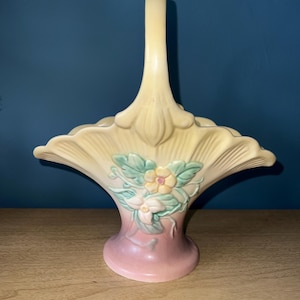 May include: A ceramic basket with a handle, featuring a light yellow top and a pink base. The basket is adorned with a floral design in shades of green, pink, and yellow. The handle is a solid, curved shape.