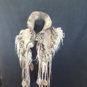 May include: A beige felted wool scarf with a large collar and long fringe. The scarf is displayed on a black stand.